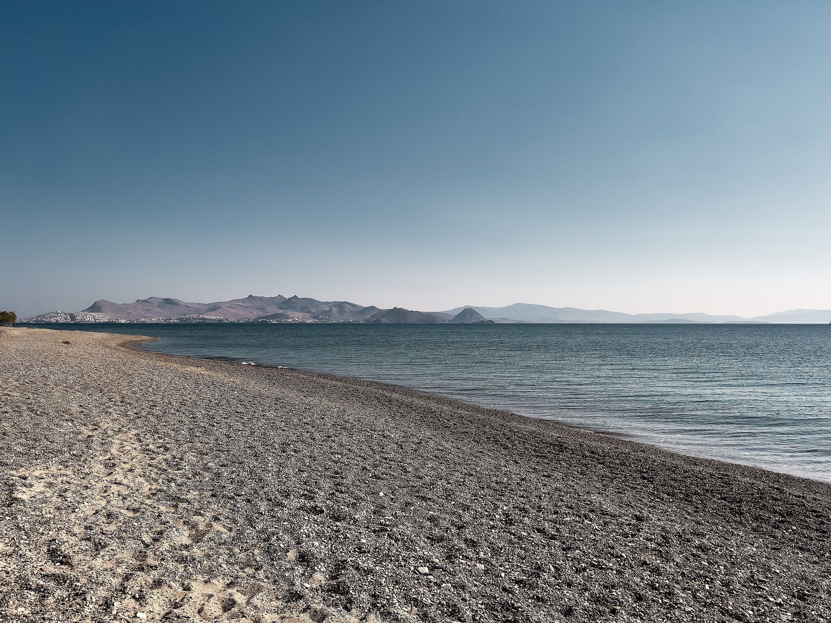 A pebble beach with a calm sea and distant mountains.