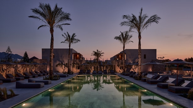 A pool area with lounge chairs and umbrellas, surrounded by palm trees and modern buildings. - Theros All Suite Hotel - Adults Only (Kos)