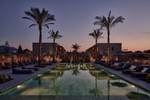 A pool area with lounge chairs and umbrellas, surrounded by palm trees and modern buildings. - Theros All Suite Hotel - Adults Only (Kos)