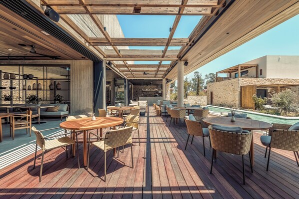 An outdoor patio with wooden tables and chairs, a pool, and a view of a modern building. - Theros All Suite Hotel - Adults Only (Kos)