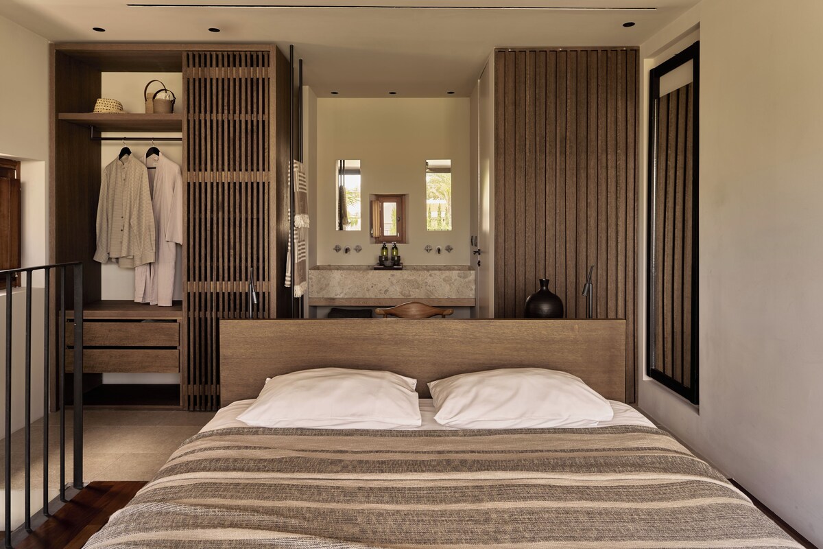 A bedroom with a large bed, a wooden headboard, a wooden wardrobe, a bathroom with a mirror, and a window.