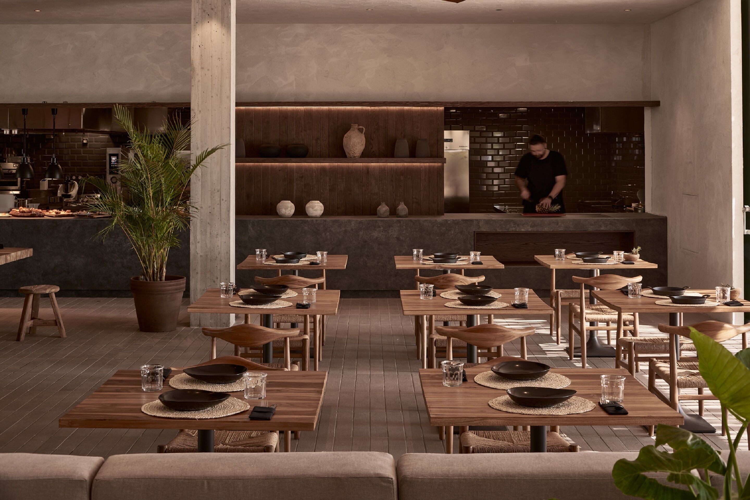 Un ristorante moderno con tavoli e sedie in legno, uno chef che prepara cibo, e una parete di mattoni sullo sfondo.