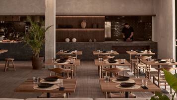 Un ristorante moderno con tavoli e sedie in legno, uno chef che prepara cibo, e una parete di mattoni sullo sfondo.