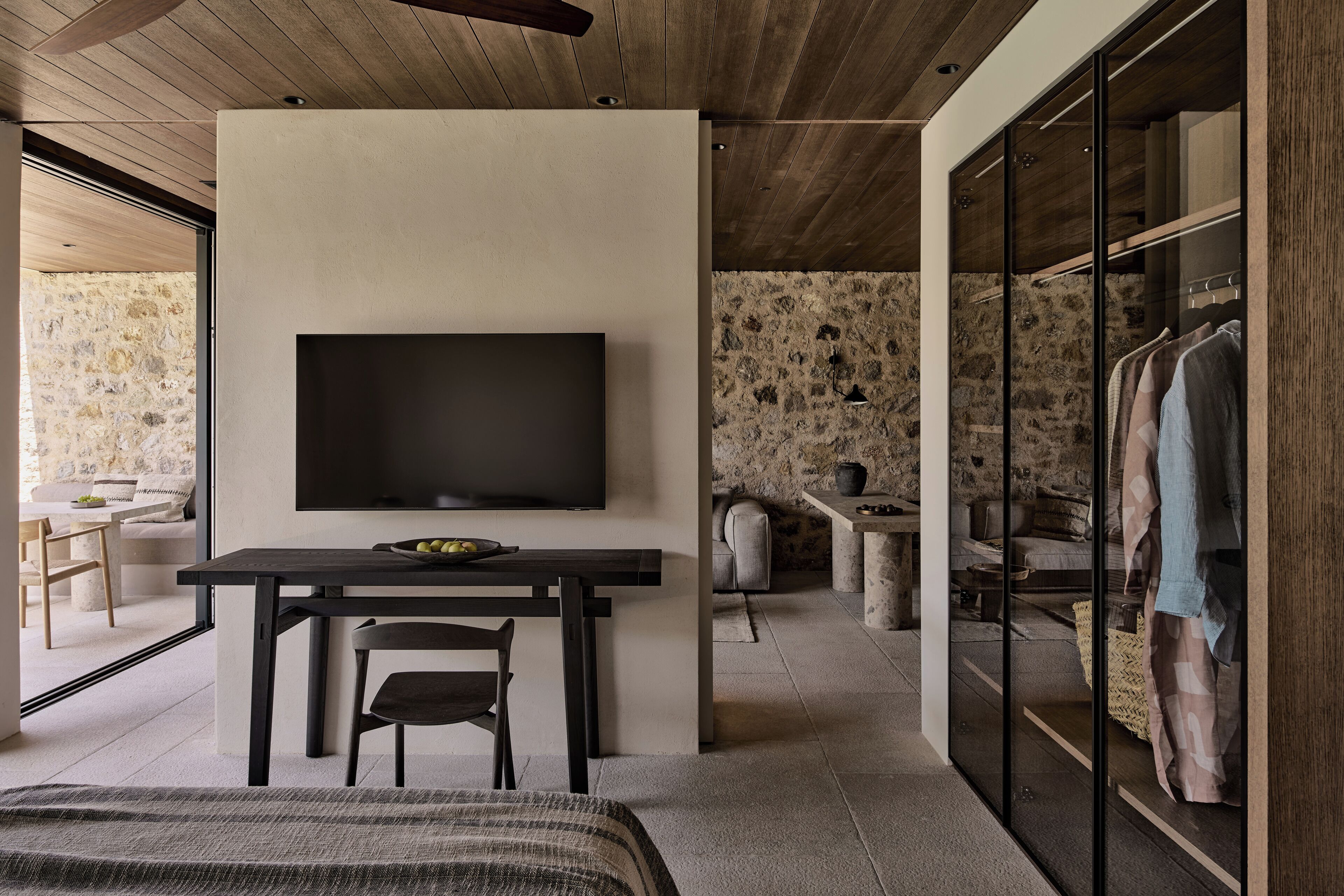 A modern bedroom with a flat-screen TV mounted on a wall, a wooden dining table, and a large window.