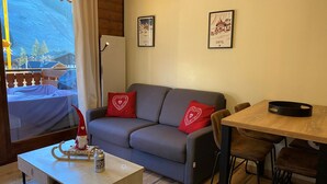 A cozy room with a grey sofa, red cushions, a wooden table, and a framed picture of a mountain town.