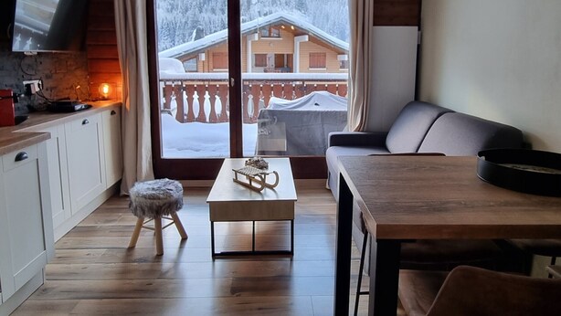 A modern living room with a wooden dining table, a sofa, and a TV mounted on the wall.