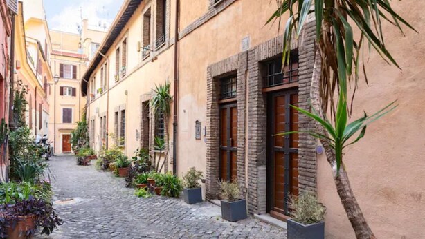 A narrow cobblestone street lined with buildings featuring traditional architecture, potted plants, and a palm tree. - Ciancaleoni Studio Atelier of the Artist - Monti (Roma)