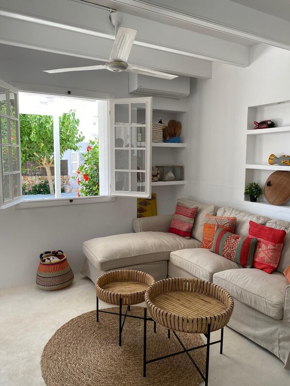 Una sala de estar moderna con un sofá blanco, dos sillones de mimbre, un ventilador de techo y un ventanal con vistas a zonas verdes.