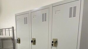 A set of grey metal lockers with ventilation slits, a metal shelf unit, and a bed with a floral bedspread.