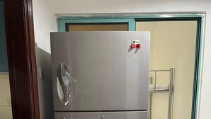 A compact, two-door refrigerator with a metallic finish, positioned in a room with tiled flooring and a wooden door frame.