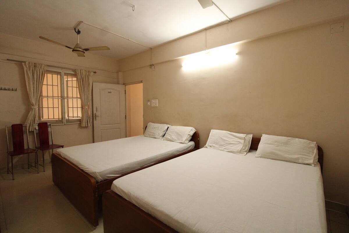 Quarto com duas camas de solteiro, um ventilador de teto, janela com cortinas e uma escrivaninha com cadeira.