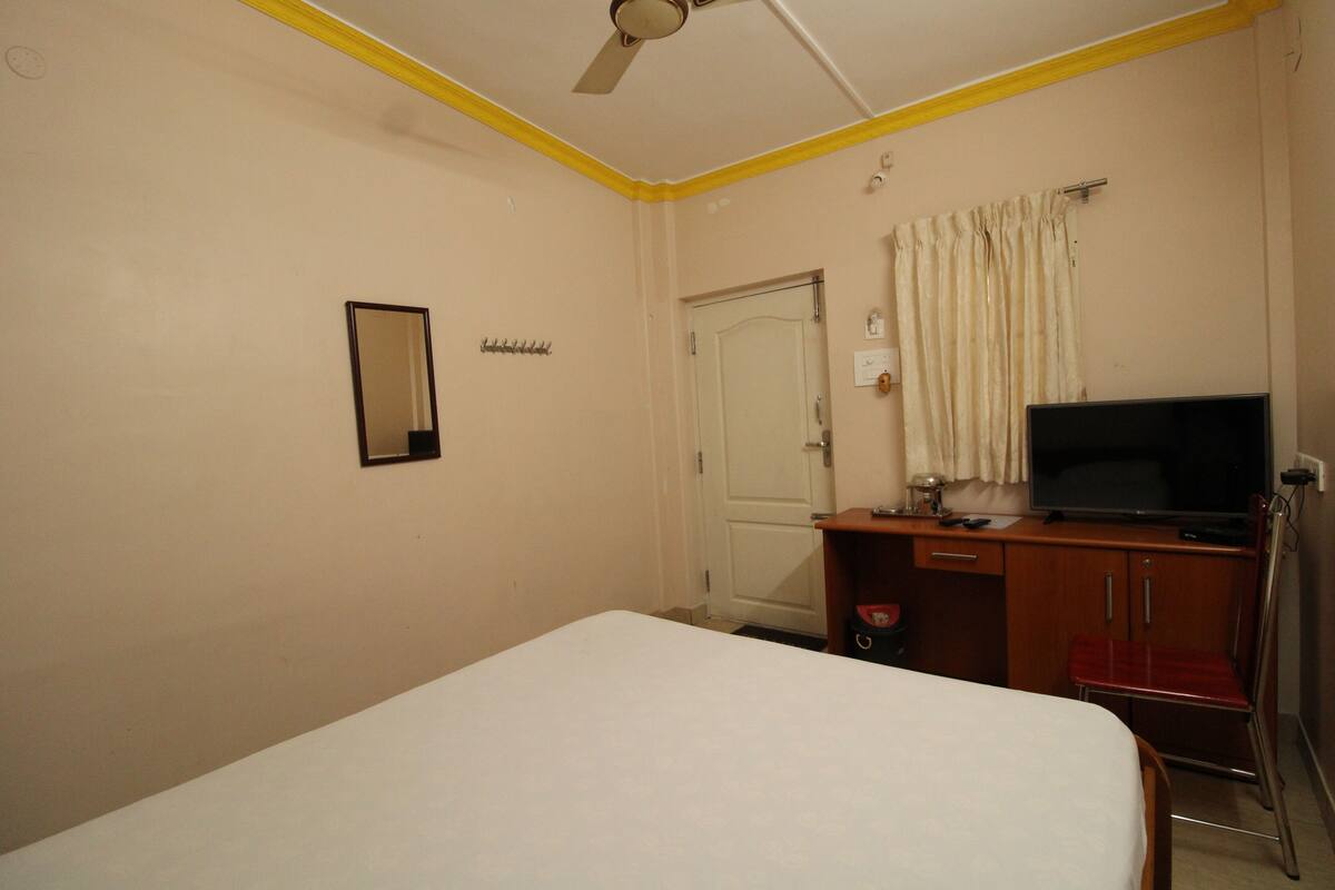 Quarto de hotel com uma cama de solteiro, televisão, escrivaninha e uma cadeira.