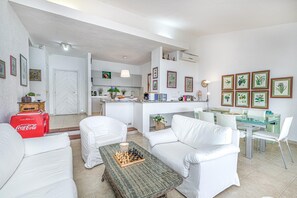 A modern living room with white furniture, a glass dining table, and a kitchen in the background.