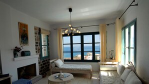 A cozy living room with a fireplace, a sofa, a small round table, and a view of the sea through large windows. - Luxury Villa with Mountain and Sea View (Siteia)