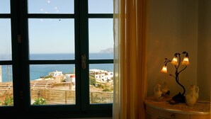 A room with a large window overlooking a coastal area, a bedside table with decorative items, and a curtain partially drawn. - Luxury Villa with Mountain and Sea View (Siteia)