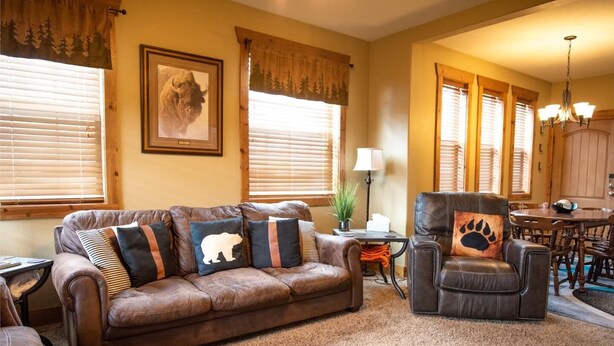 Una sala de estar acogedora con un sofá de cuero marrón, un sillón de cuero y una almohada con huella de pata, y un comedor con muebles de madera.