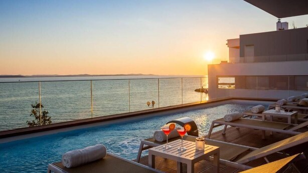Zona de piscina en la azotea con sillones reclinables y una mesa, con vistas a un cuerpo de agua al atardecer.