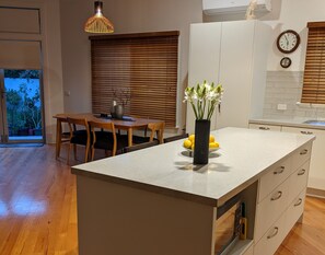 A modern kitchen with a central island, wooden dining table, and a view of the outdoors. - Foxhouse (Aireys Inlet)