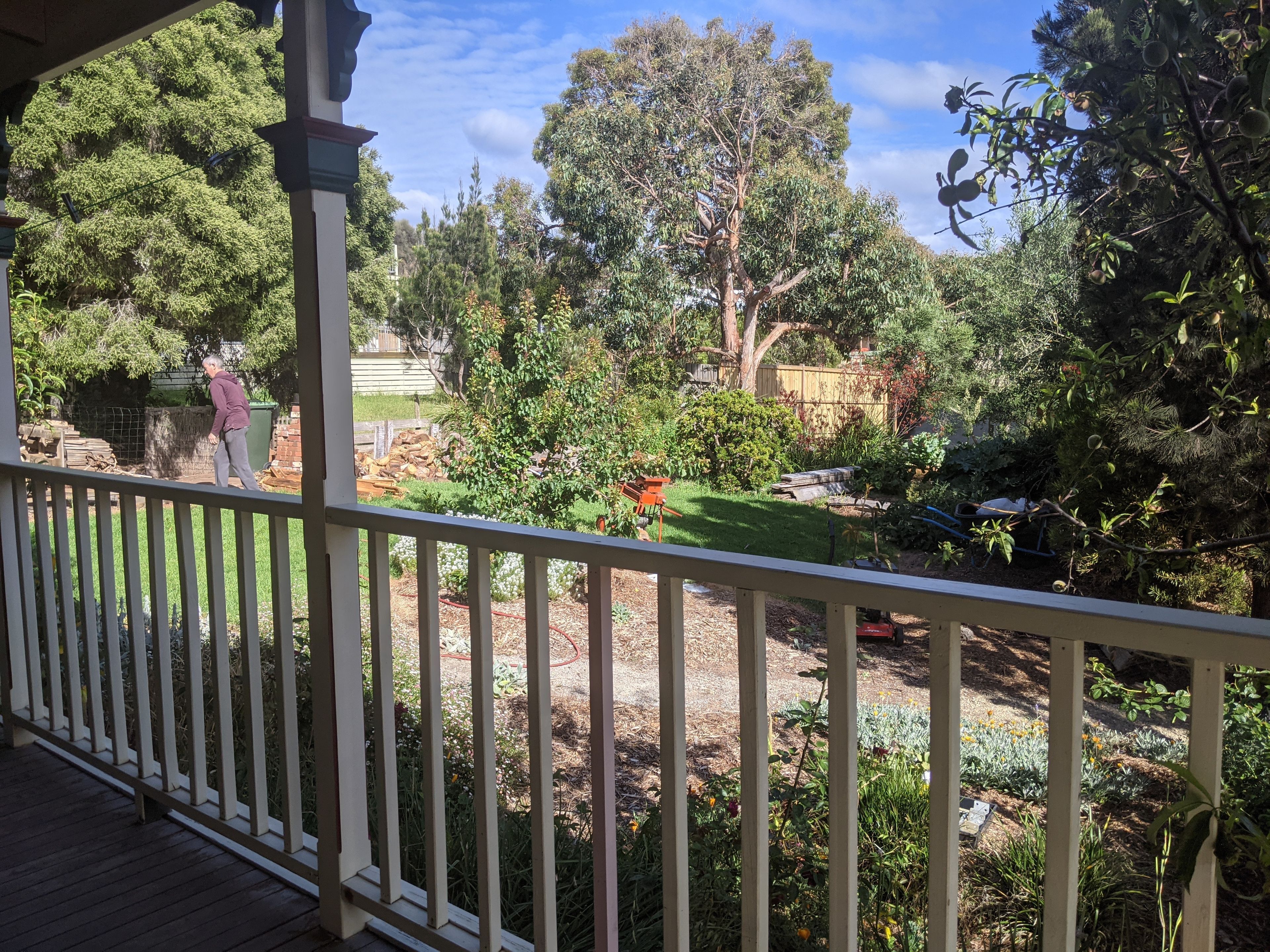 A covered outdoor area with a wooden deck, a garden with various plants and trees, and a person standing by a pile of firewood.