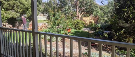 A covered outdoor area with a wooden deck, a garden with various plants and trees, and a person standing by a pile of firewood.
