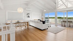 A spacious living area with a white sofa, wooden dining table, and chairs. There is a large window providing a view of the outdoors. - Peppertree Jervis Bay - Pet Friendly Beachfront with Sea Views (Callala Beach)