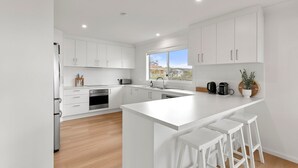 A modern kitchen with white cabinetry, a large island with bar stools, a built-in oven, and a window providing natural light. - Peppertree Jervis Bay - Pet Friendly Beachfront with Sea Views (Callala Beach)