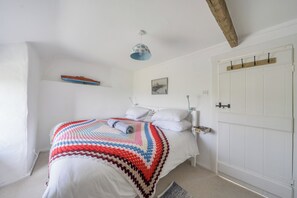 Una camera da letto con un letto, pareti bianche, travi in legno a soffitto, una lampada a sospensione, una porta con maniglia e un piccolo ripiano con un quadro.