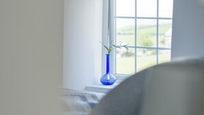 Ett fönster med utsikt över ett grönskande landskap, en blå vas med en växt på fönsterbrädan och en vit vägg.