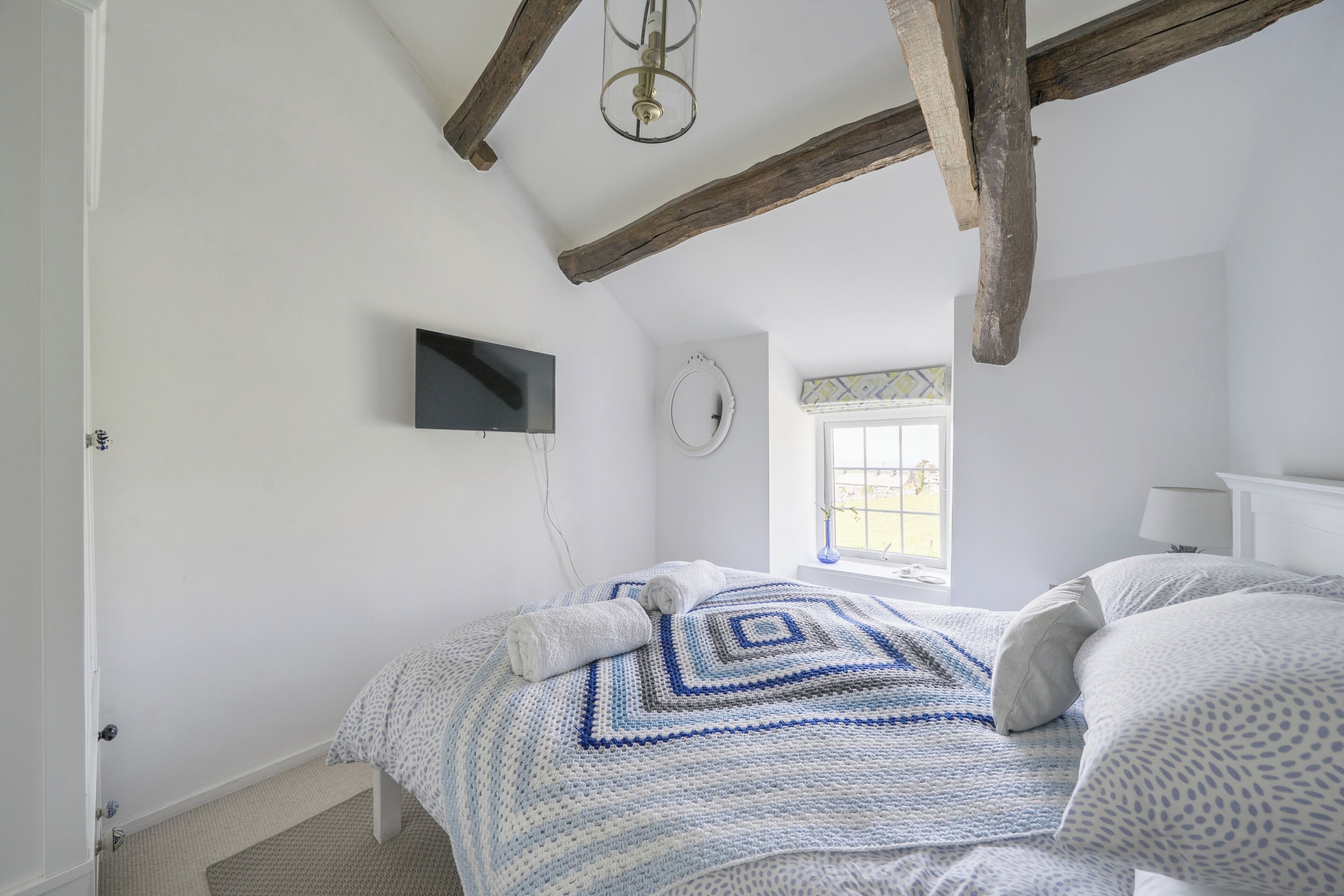 A bedroom with a bed, a television, a window, and a wooden ceiling beam.