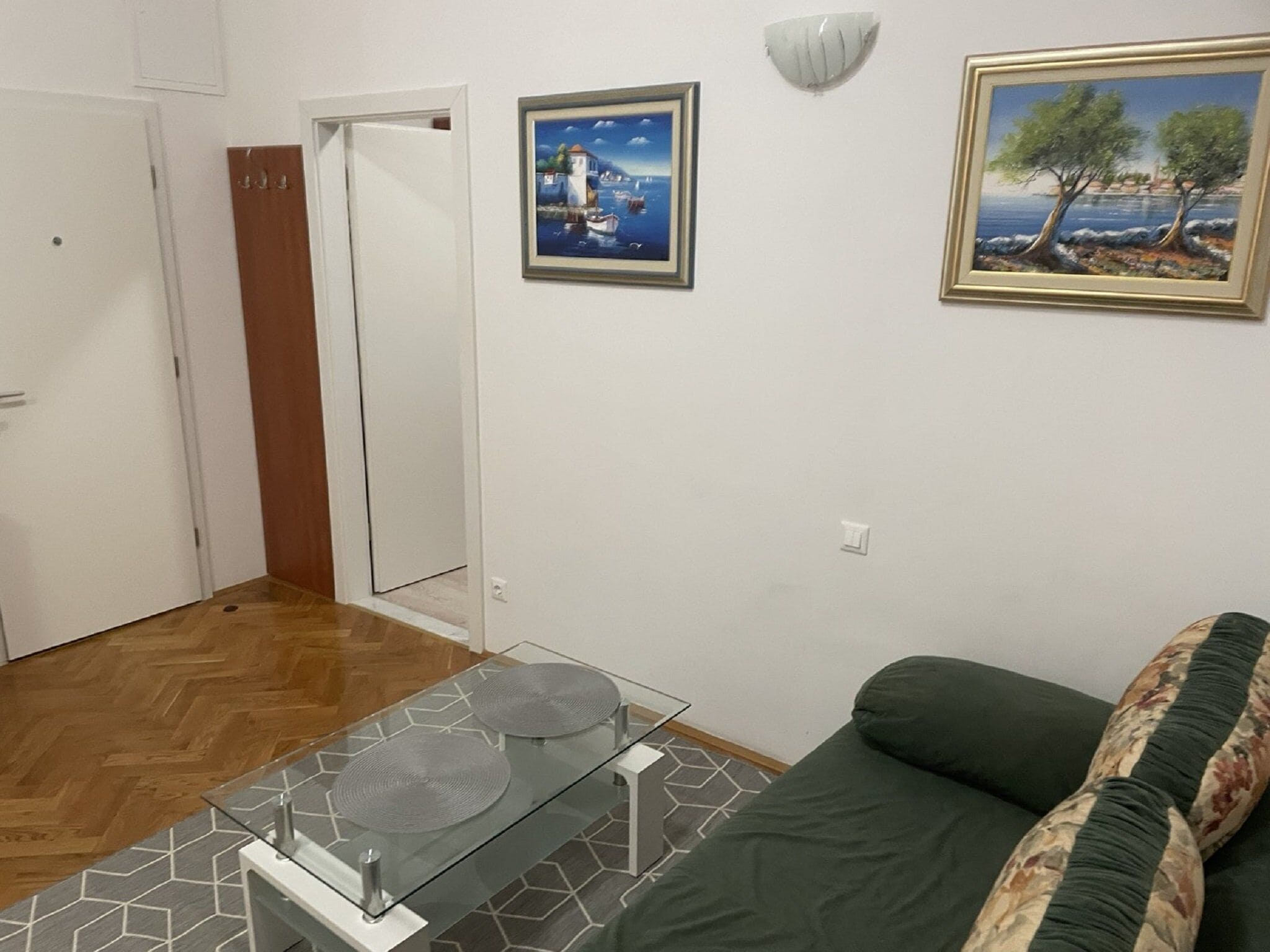 A room with a green sofa, a glass table with two circular objects, a painting of a coastal scene, and a door with a wooden panel.