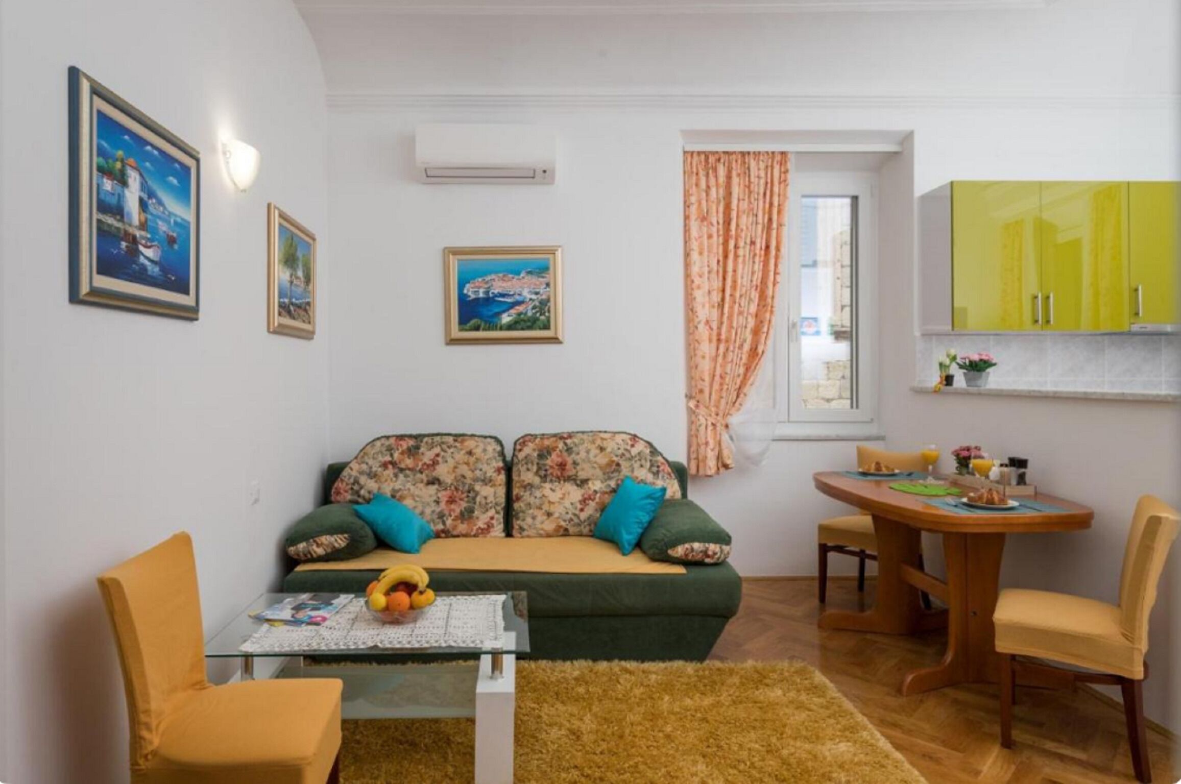 A cozy living room with a sofa, a coffee table, and a dining table with chairs. There are framed pictures on the wall, a window with curtains, and a bright yellow cabinet.