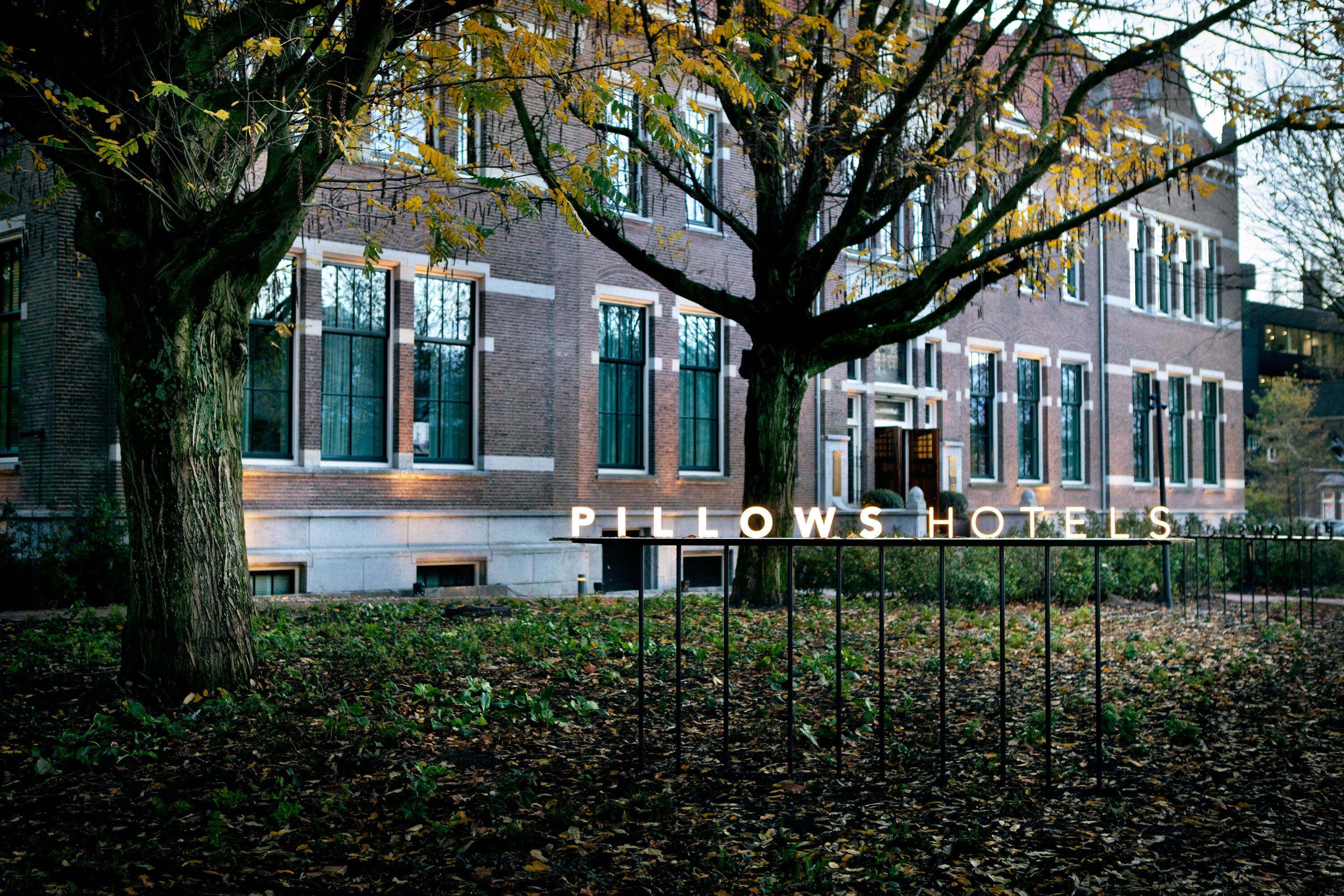 Een gebouw met een bord waarop 'PILLOWS HOTELS' staat, voor een boom en een bakstenen gevel.