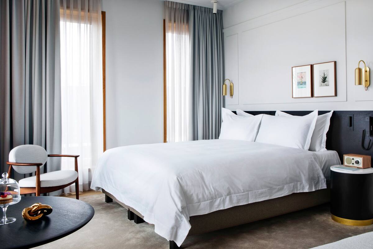 A modern hotel room with a large bed, a bedside table with a radio, a chair, and a round table with a glass dome containing a dessert.