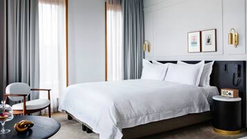 A modern hotel room with a large bed, a bedside table with a radio, a chair, and a round table with a glass dome containing a dessert.