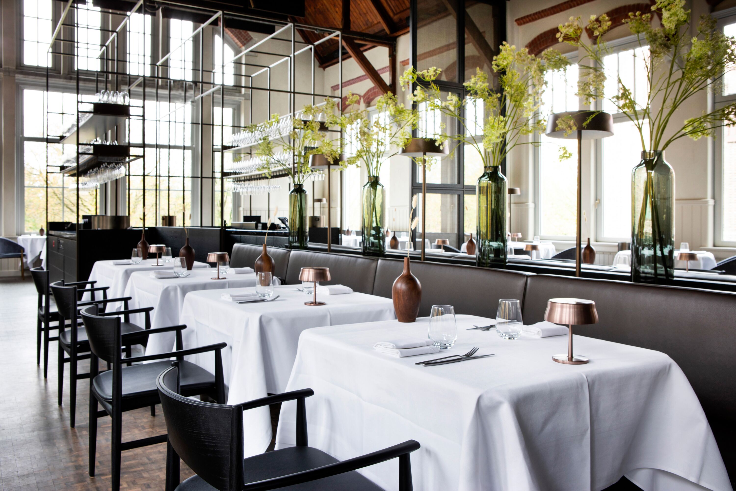 Een restaurant met witte tafellakens, zwarte stoelen en hoge vazen met bloemen.