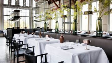 Een restaurant met witte tafellakens, zwarte stoelen en hoge vazen met bloemen.