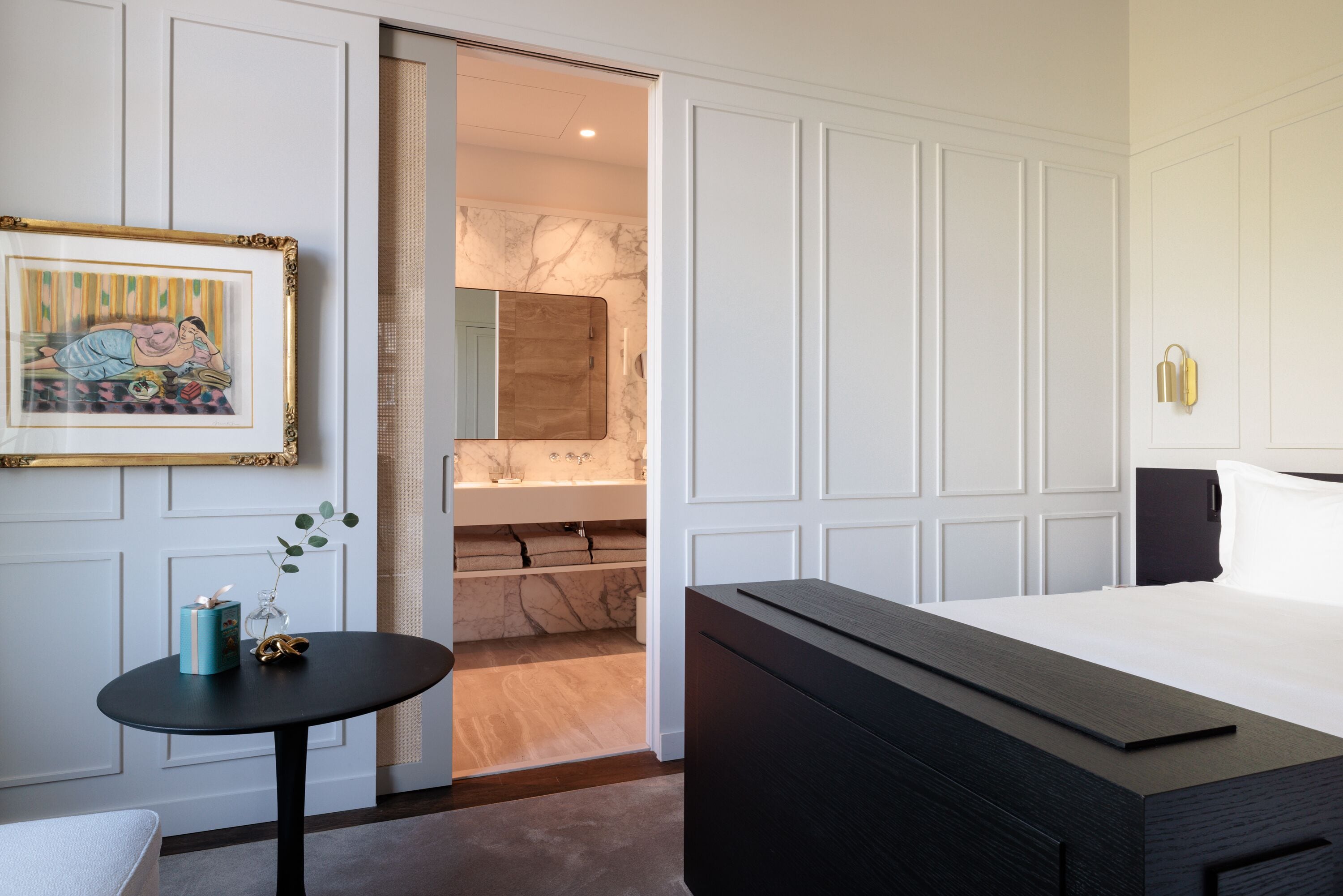 A modern hotel room with a large bed, a small table with a vase, a framed picture on the wall, and a bathroom visible through an open door.