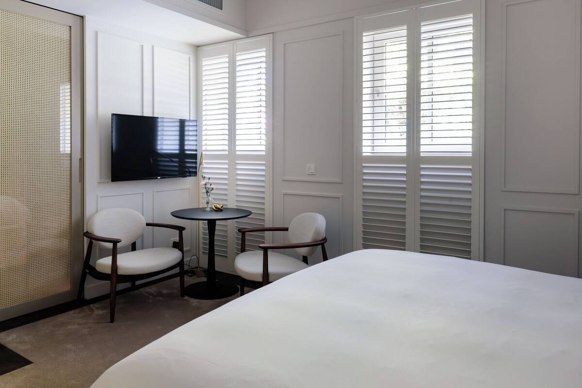A hotel room with a bed, two chairs, a small table, a TV, and large windows with blinds.