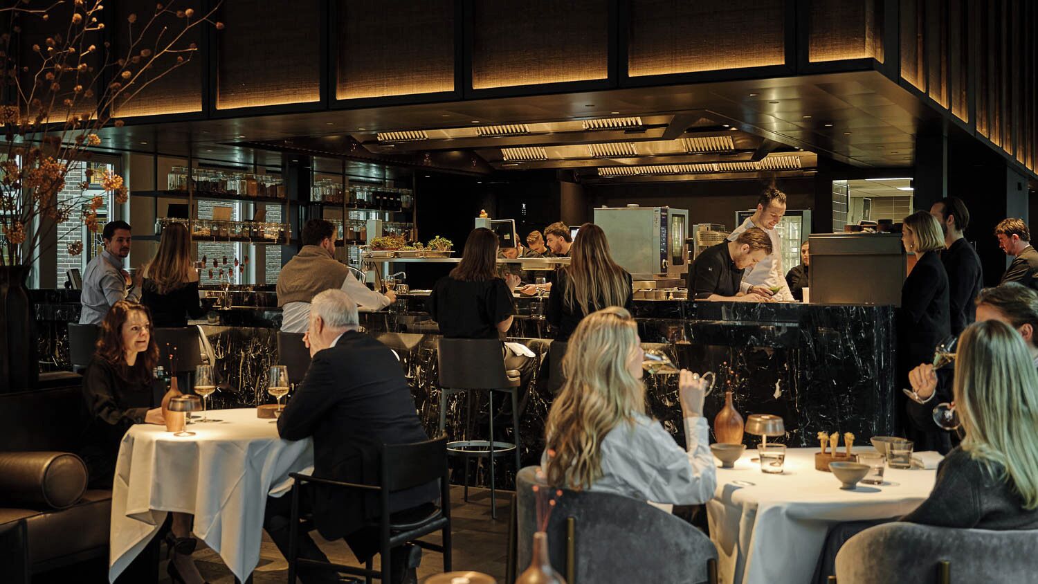 A restaurant with tables set for dining, a bar area with staff, and a kitchen in the background.