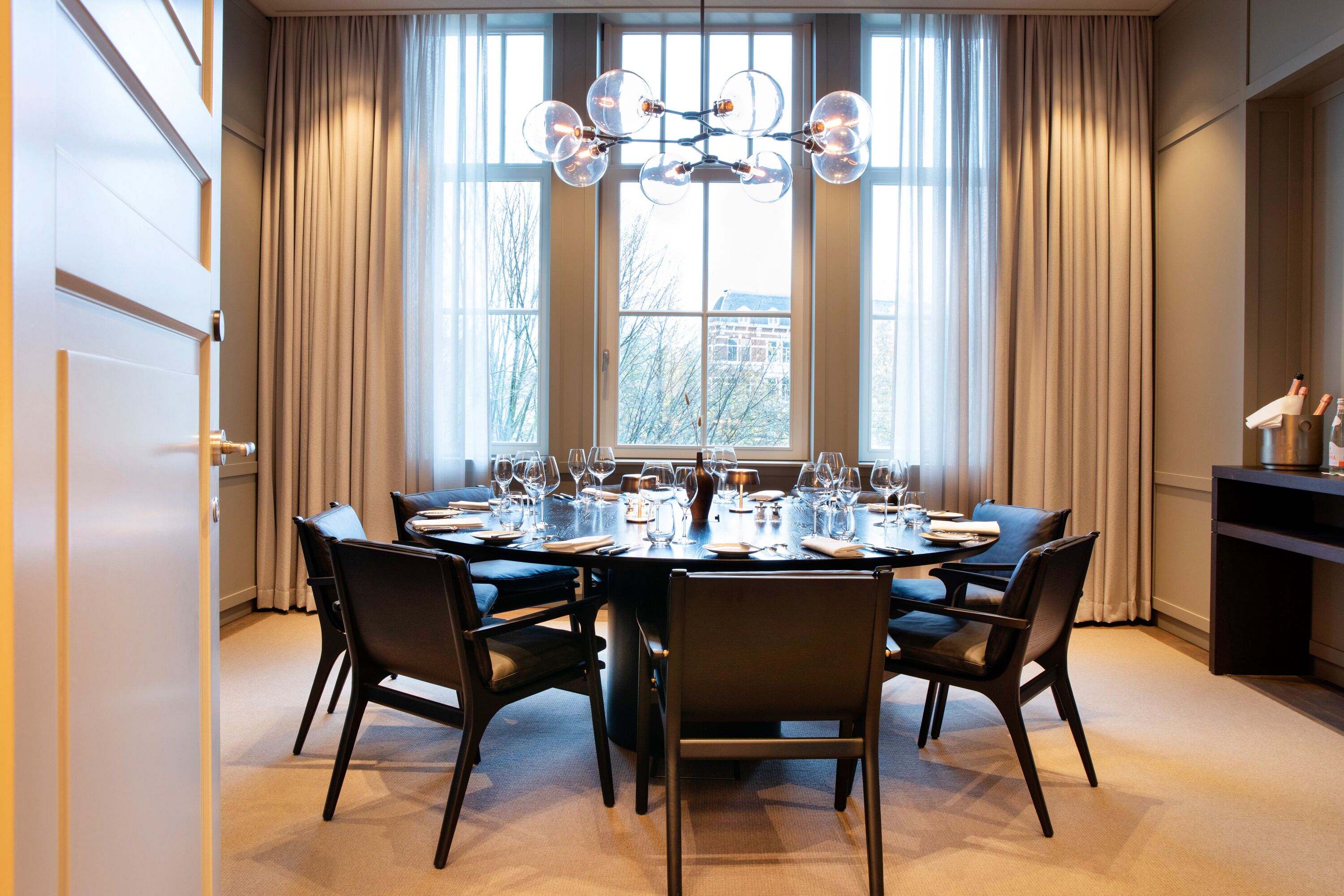 A dining area with a round table set for six, large windows with curtains, and a chandelier.