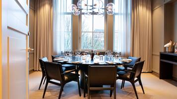 A dining area with a round table set for six, large windows with curtains, and a chandelier.