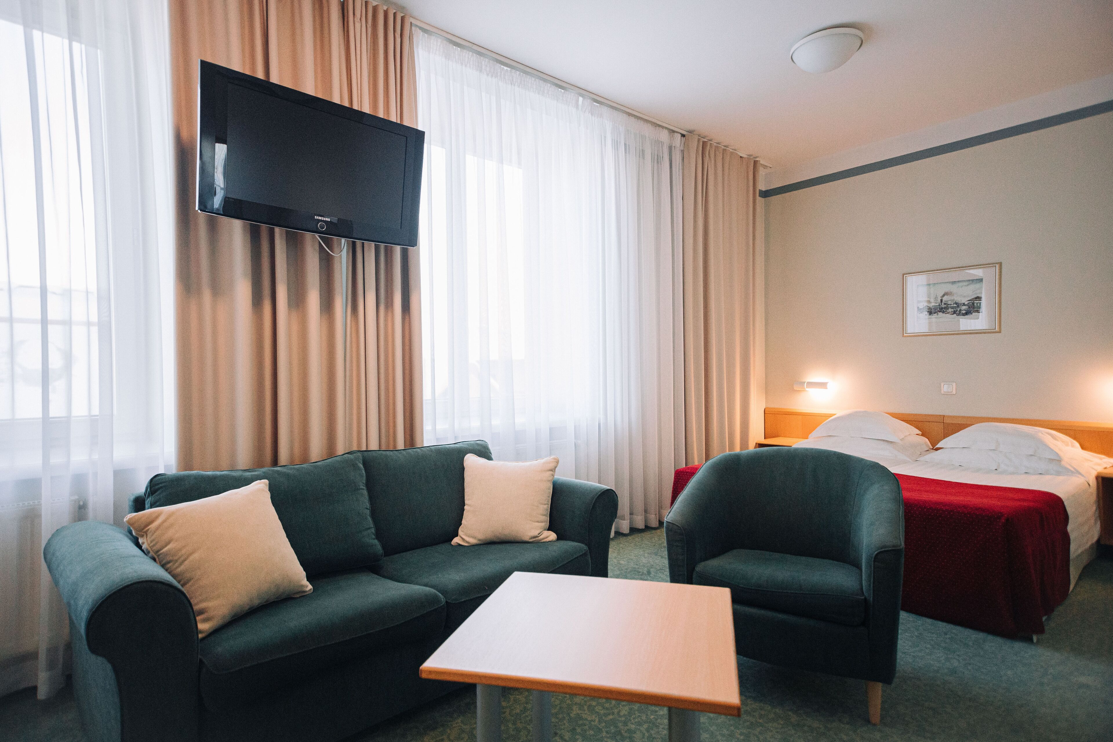 Ein Hotelzimmer mit einem an der Wand befestigten Flachbildfernseher, einem Bett mit roter Bettdecke, einem grünen Sofa, einem kleinen Tisch und einem gerahmten Bild.
