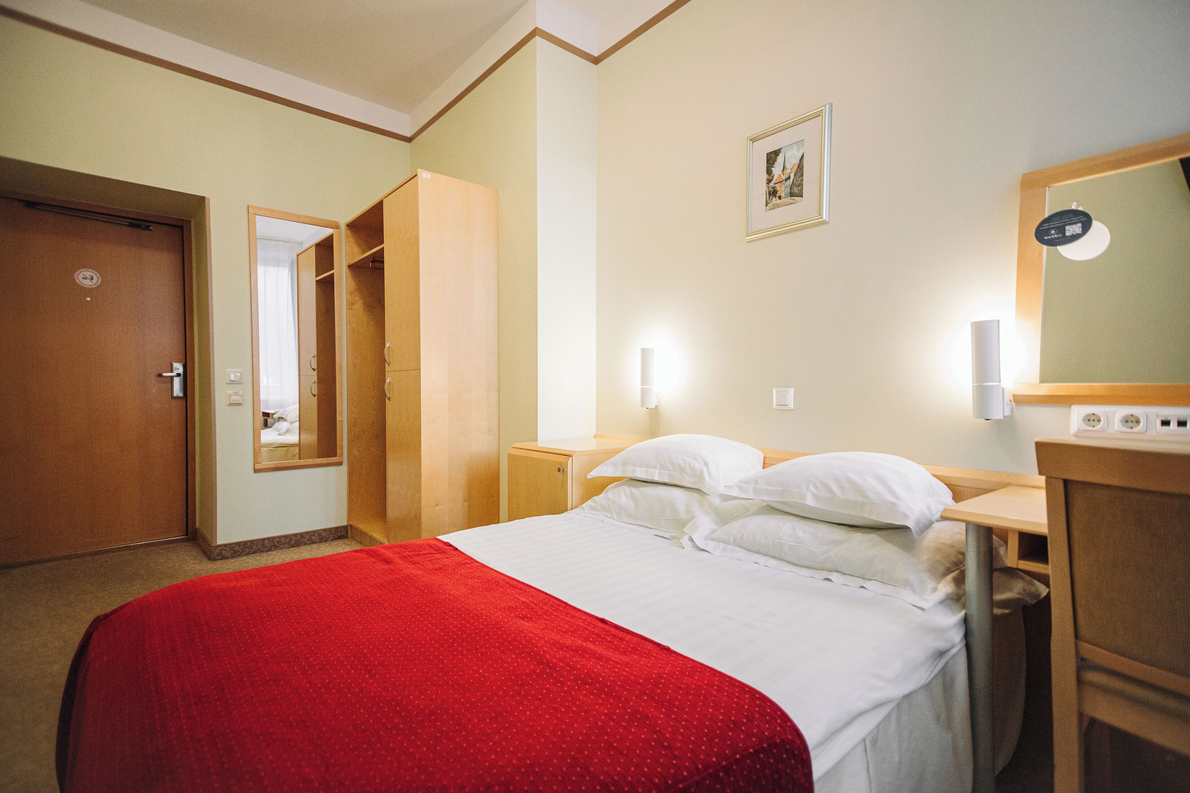A hotel room with a bed, a red blanket, a wooden wardrobe, a bedside table, a nightstand, a mirror, and a wall-mounted light fixture.