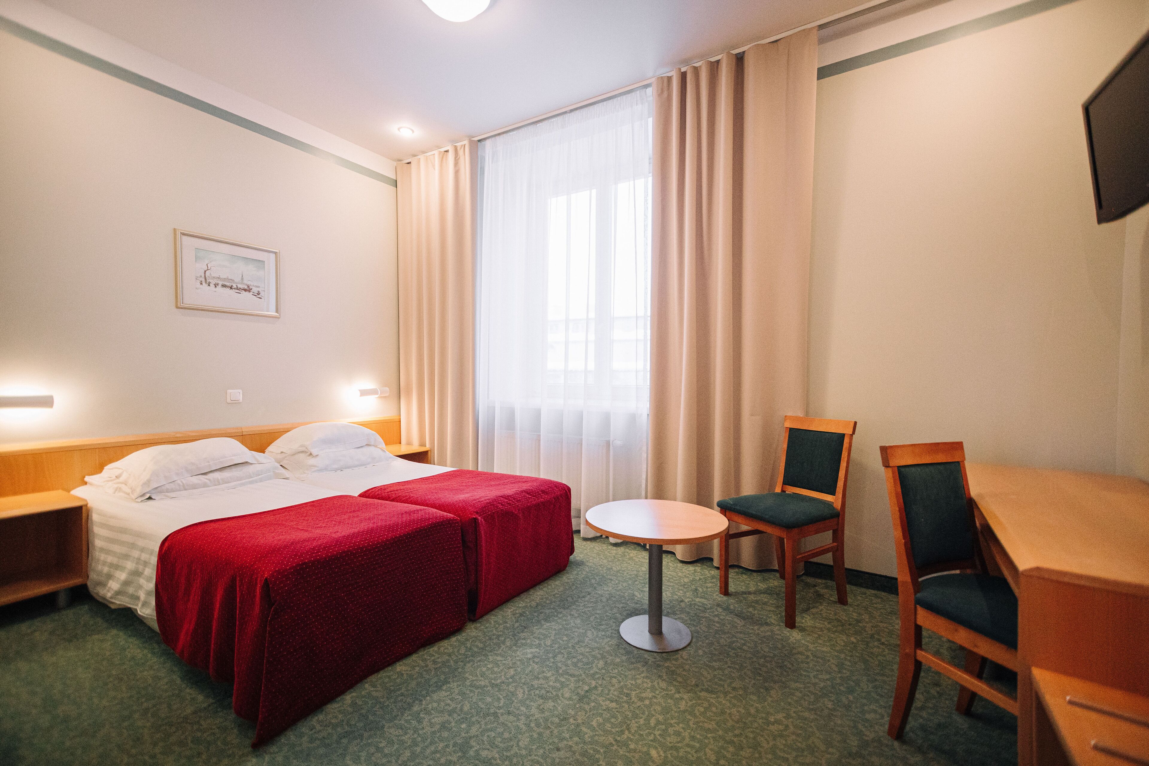 A hotel room with a bed, two red bedsheets, a small round table, two chairs, a television, and a window with curtains.