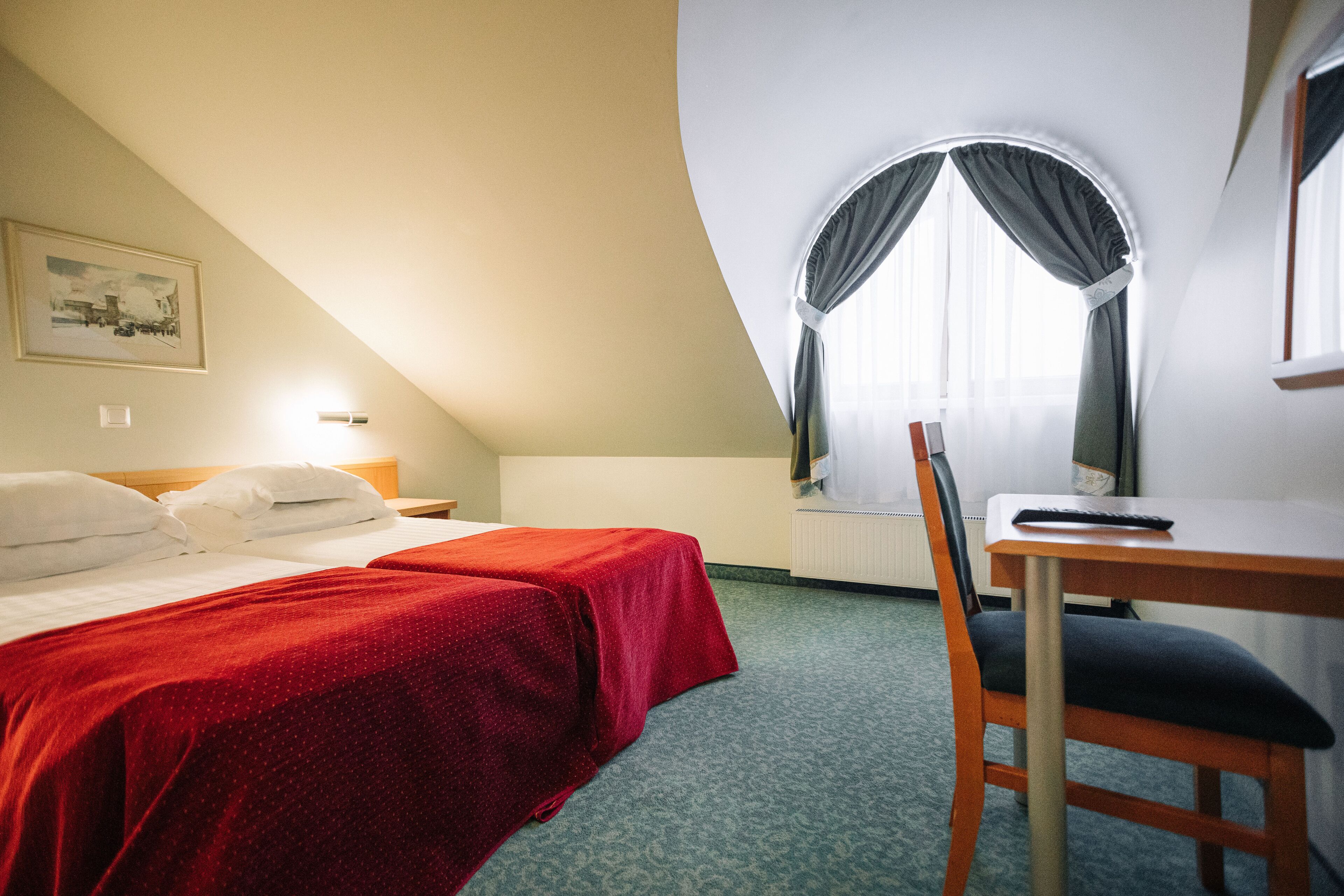 A hotel room with a bed, a desk, a chair, a window with curtains, a radiator, and a painting on the wall.