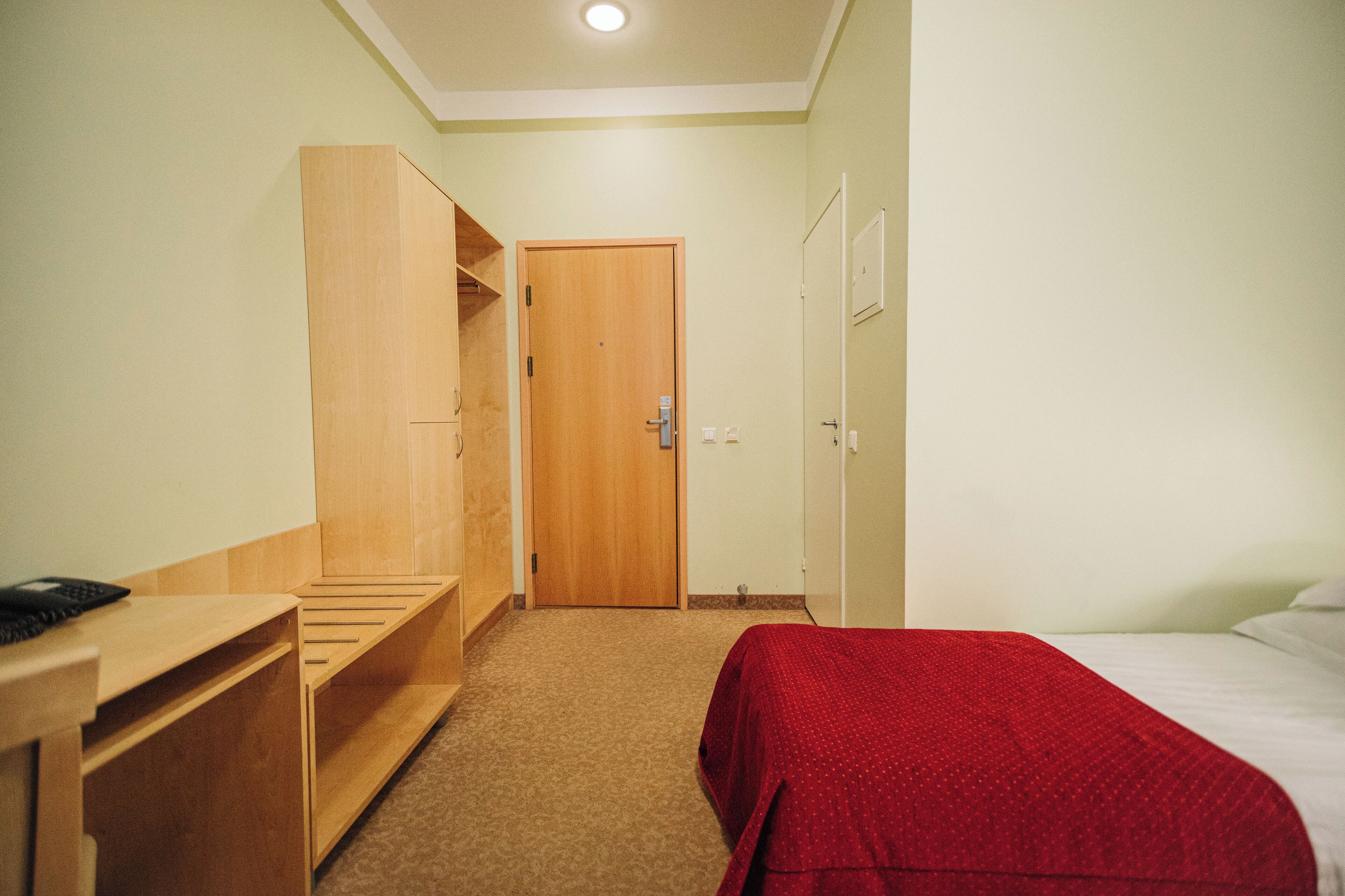 A small, neatly organized bedroom with a wooden wardrobe, a bed with a red blanket, and a desk with a telephone.