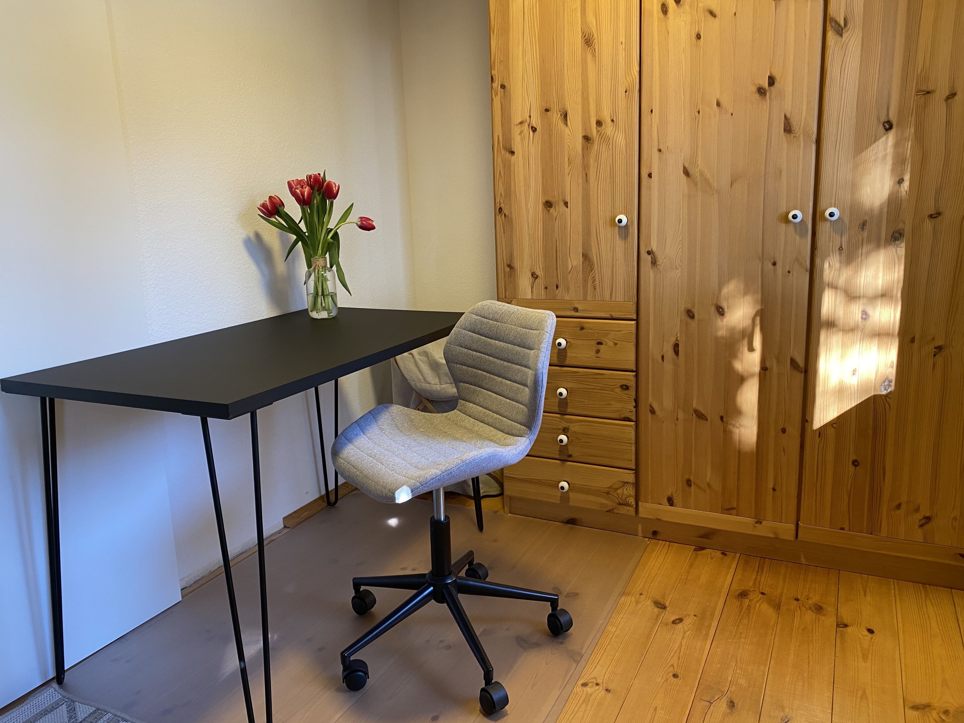 Een werkplek met een zwart bureau, een grijze bureaustoel en een vaas met rode bloemen.