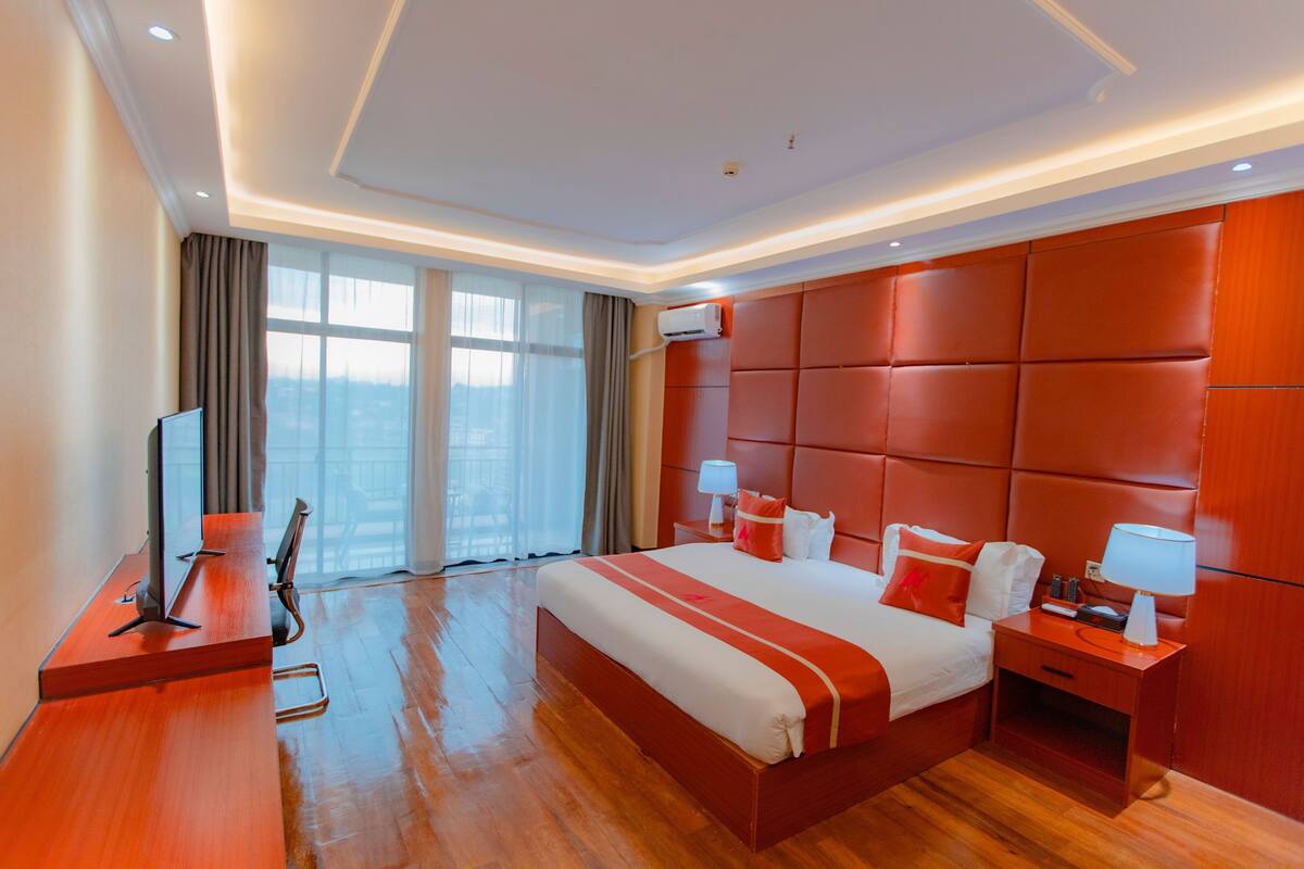 A hotel room with a large bed, wooden floor, a TV, a desk, and a window with curtains.
