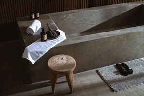 Une salle de bain avec un lavabo en marbre, un tabouret en bois, une serviette, une bouteille et une paire de chaussures posées sur un tapis texturé.