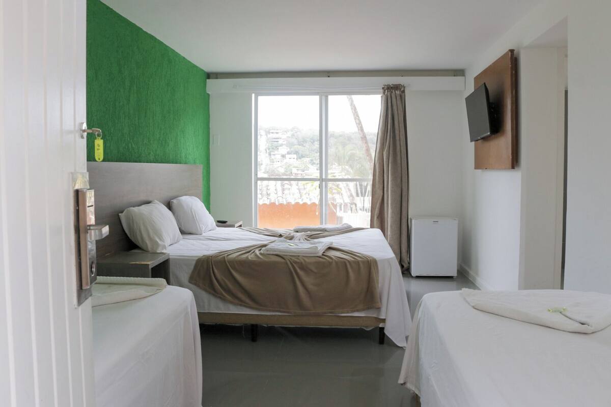 A hotel room with a bed, a small bedside table, a TV mounted on the wall, and a window with curtains.